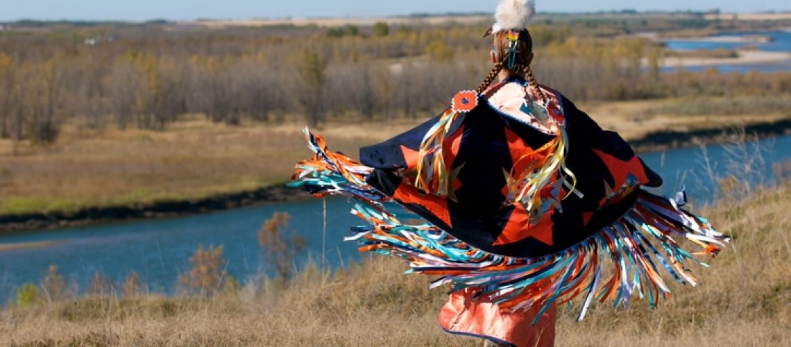 A woman performing a First Nations fancy shawl dance in a field alongside the river in Saskatoon, Saskatchewan