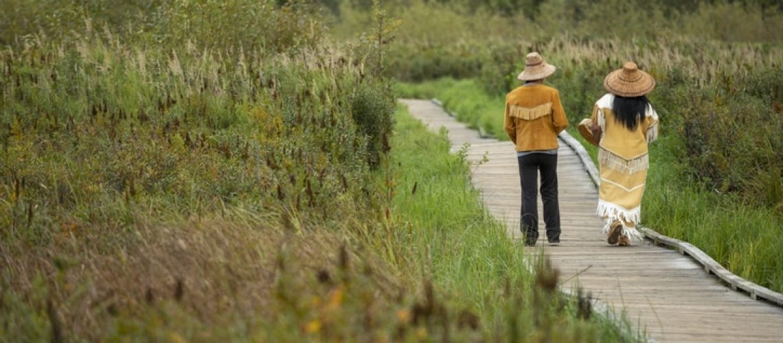Indigenous women wearing traditional outfits walks down boardwalk over marsh together